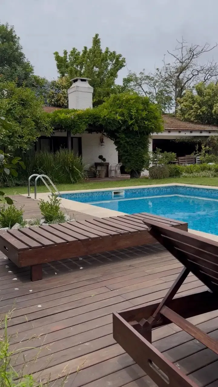 Piscina con deck de madera y reposeras, césped y una casa blanca cubierta de enredadera al fondo